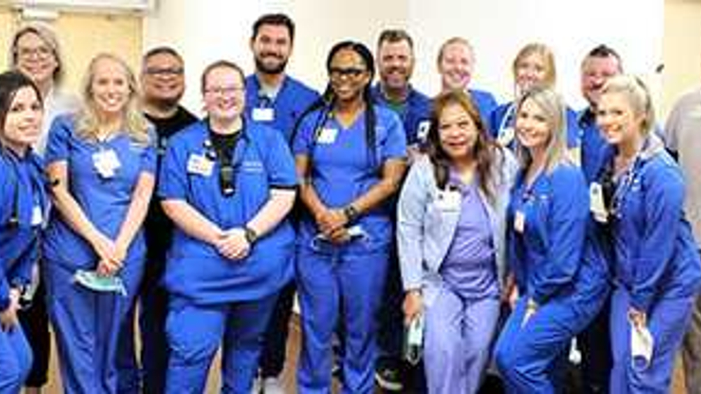 A group of female and male nurses dressed in hospital scrubs smiling in hospital hallway.
